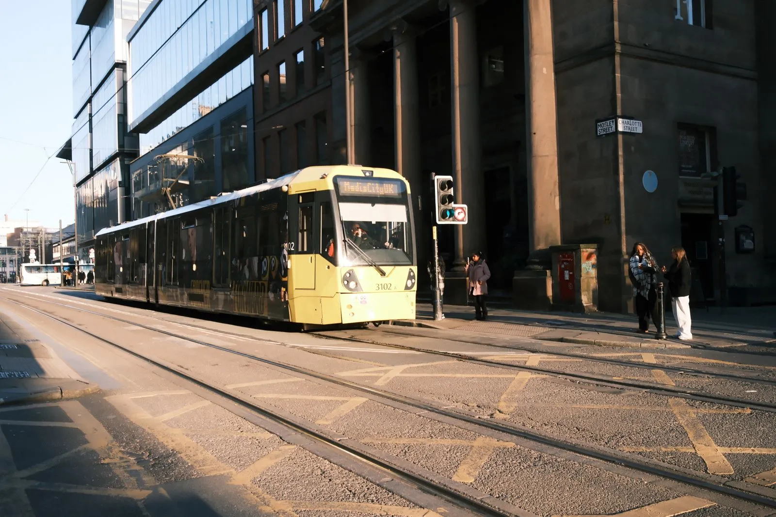 A tram travels through Manchester's bustling city streets under clear skies.